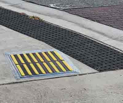 Image of a intersection corner.  Close up of Tactile Direction Indicator (2’x2’ square with raised yellow bars against a black background) placed parallel to traffic to indicate the correct alignment.  The TDI is placed on the sidewalk in front of a blended, rounded corner with black truncated domes.